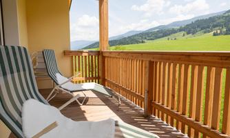 A cozy balcony with lounge chairs and a view of green meadows and mountains. The sun is shining and the sky is blue.