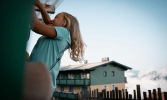A girl with long hair is standing under a roof and looking up. In the background, there is a building with balconies and a beautiful sky.