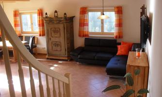 A cozy living room with a black sofa and a wooden cabinet. Bright windows with orange curtains and a small table complete the space.