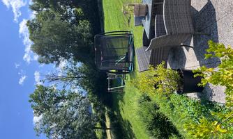 A beautiful terrace with garden furniture and a plant pot. In the background, you can see a meadow with a trampoline and trees.