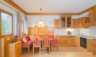 A cozy kitchen with a wooden dining area and checked cushions. The room is bright and inviting, with large windows and modern appliances.