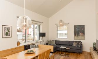 A modern living room with high ceilings and large windows. There is a wooden dining area and a cozy seating arrangement with a minimalist design.
