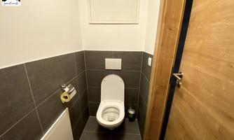 A modern bathroom with a clean toilet and dark tiles. The walls are light, and the wooden door frame adds a warm accent.