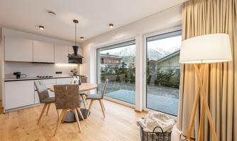 A modern kitchen with a dining area and large windows. Outside, there is a quiet garden and the mountains in the background.