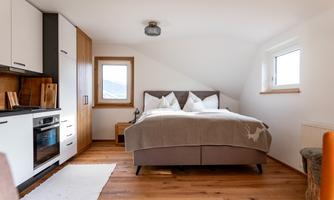 A modern bedroom with a comfortable bed and a small kitchenette. The room has light walls and large windows that let in a lot of light.