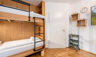 A modern room with a wooden bunk bed and warm wooden flooring. There is a television and shelves on the wall.