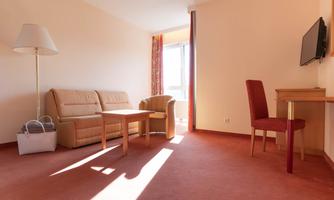 A bright room with a sofa, a chair, and a table. A television hangs on the wall, and daylight streams through the window.