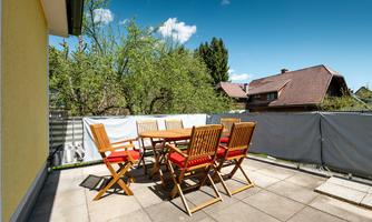 A cozy terrace area with a wooden sign and chairs. The space is sunny and surrounded by green trees.