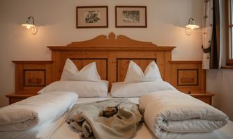 A cozy bedroom with a wooden bed and white pillows. On the bed are a blanket and a small jug.