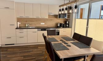 A modern kitchen with wooden flooring and bright furniture. The dining table is set with placemats and there are large windows for plenty of natural light.
