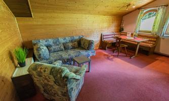 A cozy room with a wooden panel ceiling, a sofa, and an armchair. A table with chairs and a window provide light and an inviting atmosphere.