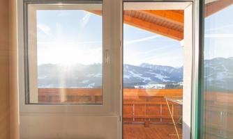 A bright room with an open window overlooking a terrace and the mountains. The sun is shining and the sky is clear.