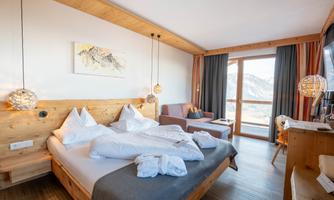A modern hotel room with wooden decor, a large bed, and a cozy seating area. Large windows provide a beautiful view of the mountains.