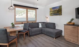 A modern living space with a gray sofa and a wooden table. Large windows offer a view of nature, and a painting complements the cozy atmosphere.