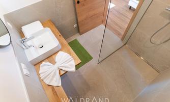 A modern bathroom with an elegant wooden vanity top and a rectangular sink. In the shower area, there is a green bath mat and an artfully folded towel.