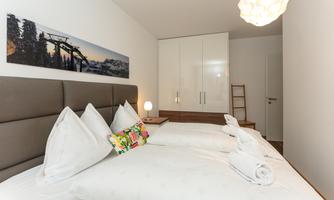 A cozy bedroom with two beds and large, white pillows. The walls are bright and a modern lamp provides pleasant light.