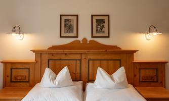 A cozy bedroom with two single beds, surrounded by wooden furniture. Above the beds hang two pictures and there are lights mounted on the walls.