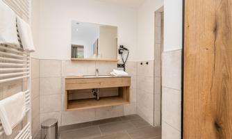 A modern bathroom with a wooden vanity and a large mirror. The walls are tiled in light colors and there are towels on a radiator.