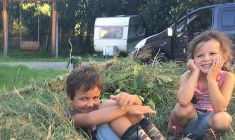Two children are playing in a pile of hay. In the background, a caravan and a car can be seen.