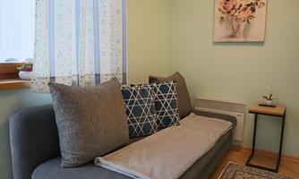 A cozy living room with a gray sofa and decorative pillows. A floral painting hangs on the wall, and a small table stands next to it.