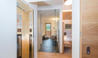 A modern hallway with a glass door and wooden floor. In the background, a bedroom with a balcony is visible.