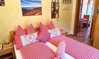 A cozy bedroom with red checkered patterns and heart-shaped pillows. In the background, a table and a door to another room are visible.