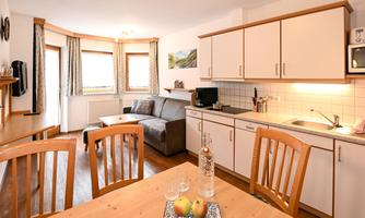 A modern kitchen with a dining table and chairs. In the background, there is a cozy living room with a sofa.