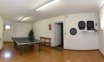 A room with a table tennis table and a dartboard on the wall. The floor is wooden, and there is a bench to sit on.