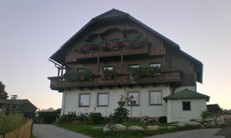 A traditional wooden house with flower balconies and a small outbuilding. Surrounded by green grass and trees.