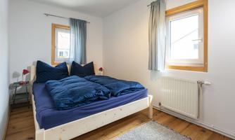 A cozy bedroom with a large bed and blue bed linen. The room has two windows and a bright wooden floor.