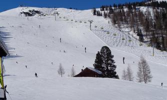 A snow-covered slope with many skiers. In the background, mountains and a blue sky can be seen.