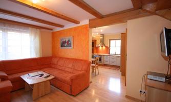 A cozy living room with an orange couch and a wooden table. The open kitchen is visible in the background, illuminated by natural light.