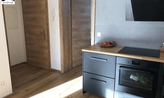 A modern kitchen with wooden flooring and dark cabinets. Light streams through the space, creating an inviting atmosphere.