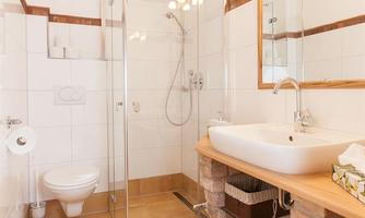 A modern bathroom with a glass shower enclosure and a sink on a wooden shelf. The walls are tiled in white and the floor is covered with terracotta tiles.