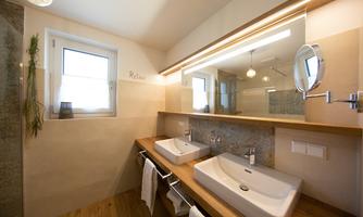 A modern bathroom with two sinks and wooden shelves. It has large windows and a bright, welcoming atmosphere.