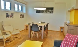 A modern dining room with wooden flooring and a large table. There are comfortable chairs and a bright ceiling lamp that makes the room inviting.