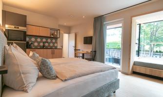 A modern room with a large bed and a small kitchenette. Large windows provide plenty of daylight and a view outside.