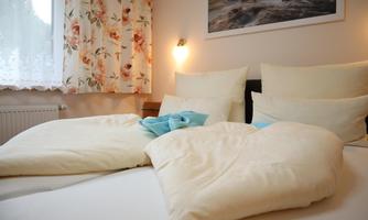 A cozy bedroom with two beds and soft, bright blankets. Gentle light and floral curtains create an inviting atmosphere.