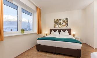 A modern bedroom with a large window that offers a view of the mountains. The decor is bright and friendly, featuring a double bed and minimalist decoration.