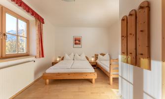 A simple and bright bedroom with wooden furniture. There is a large bed, a single bed, and a window with a view of nature.