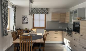 A modern kitchen with a wooden table and chairs. Bright walls and large windows provide plenty of light.