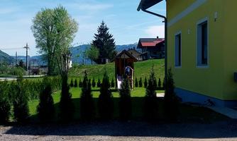 A bright yellow house stands next to a well-kept garden with shrubs. In the background, there are trees and hills, and the sky is clear.
