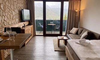 A modern hotel room with a cozy sofa and a TV. Large windows offer a view of the mountains and a balcony.