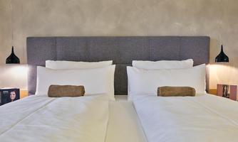 A modern bedroom with two single beds and one lamp for each. The walls are painted in neutral colors, and there are towels laid out on the beds.