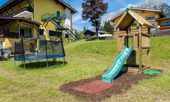 A playground with a slide and a trampoline. In the background, there are yellow houses and a green meadow.