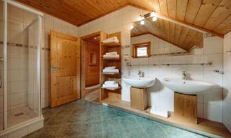A modern bathroom with a shower, two sinks, and wooden shelves. The room has a bright and inviting atmosphere due to the wooden walls and large windows.