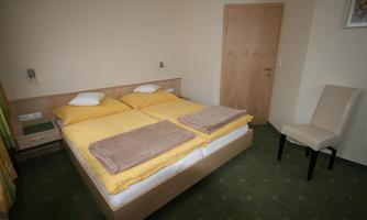 A simple bedroom with a double bed and fresh yellow bed covers. There is a chair and a wooden wardrobe door in the background.