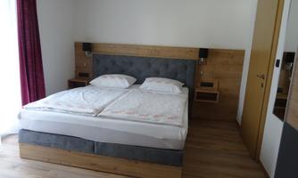 A modern bedroom with a double bed and wood-colored nightstands. Large windows with red curtains provide plenty of daylight.