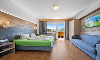 A modern hotel room with a large bed and a sofa. The room has daylight and a balcony with a view of the landscape.