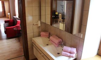 A modern bathroom with a white sink and pink towels. In the background, a cozy living area and a table are visible.